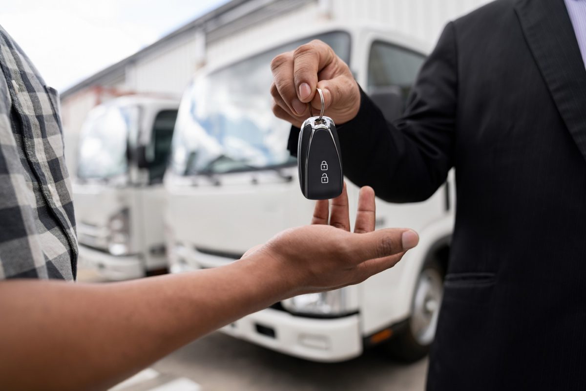 Uma pessoa entrega a chave de um veículo a outra, com caminhões brancos ao fundo.